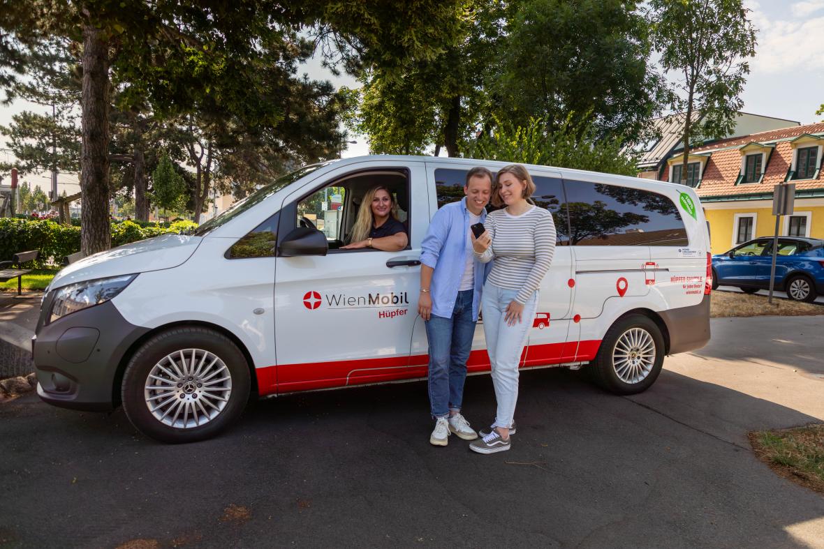 WienMobil Hüpfer: weißer Kleinbus, davor stehen zwei junge Personen und buchen via Smartphone App den Shuttleservice