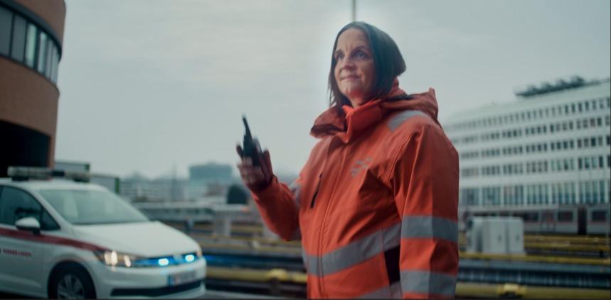 eine Frau in Sicherheitsjacke steht neben einem Einsatzwagen bei einer U-Bahn-Station und hält ein Funkgerät zum Mund