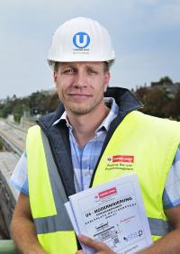 ein Mann mit einem weißen Helm mit U4 Logo und Warnweste hält Papiere in der Hand und blickt in die Kamera