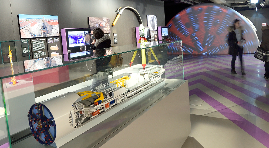 Besucherinnen im Infocenter betrachten Ausstellungsgegenstände im Vordergrund das Modell einer Tunnelvortriebsmaschine